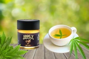 A jar of Holy Herbajuana Buzz cannabis-infused honey sits on a wooden surface next to a white teacup filled with herbal tea, garnished with a fresh cannabis leaf. The background is blurred green foliage, creating a natural and calming atmosphere.