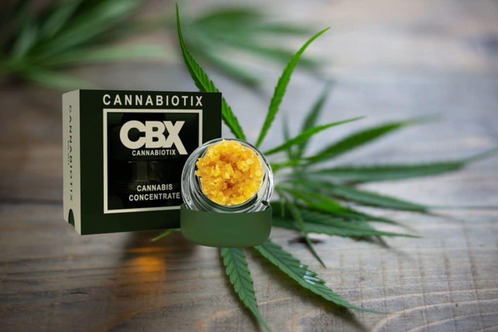 CBX Cannabiotix cannabis concentrate in a glass jar with golden extract, placed next to its black packaging and fresh cannabis leaves on a wooden surface.