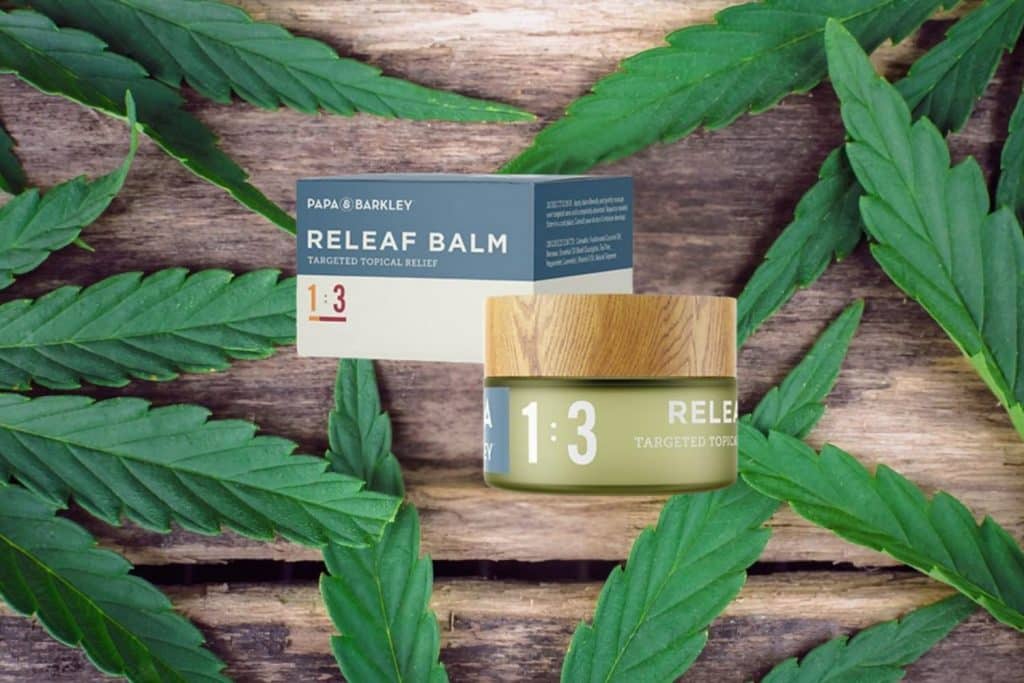 Papa & Barkley Releaf Balm 1:3 in a wooden-lid jar with matching packaging, placed on a rustic wooden surface with fresh cannabis leaves.