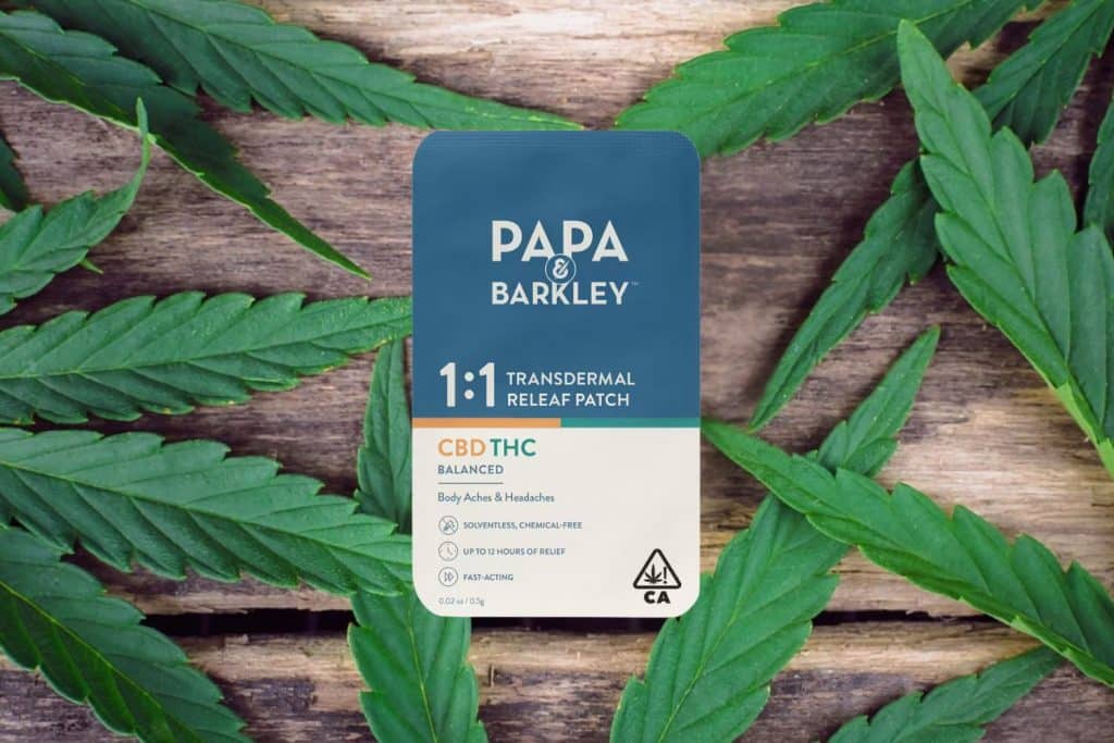 Papa & Barkley 1:1 Transdermal Releaf Patch with CBD and THC, placed on a wooden surface surrounded by fresh cannabis leaves.